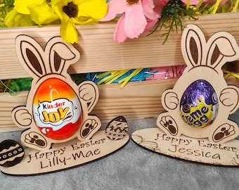 Puede incluir: Dos soportes para huevos de Pascua de madera con forma de conejo. El soporte de la izquierda tiene un huevo Kinder Joy y dice "Feliz Pascua Lilly-Mae". El soporte de la derecha tiene un huevo Cadbury Creme y dice "Feliz Pascua Jessica".