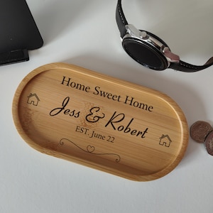 May include: An oval bamboo tray with engraved text reading "Home Sweet Home", "Jess & Robert EST. June 22", house icons, and a decorative flourish. A watch and coins are visible. The tray is light brown in colour.