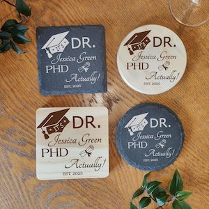Könnte beinhalten: Vier Untersetzer mit einem Abschluss-Thema. Zwei sind quadratisch und zwei sind rund. Jeder Untersetzer hat den Text "IT'S DR. Jessica Green PHD Actually! EST. 2025" mit einer Abschlusskappe und einer Diplomgrafik. Die Untersetzer sind aus Holz und Schiefer.