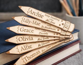 May include: Wooden pencil-shaped name tags with engraved names and messages. The tags are arranged in a fan shape, resting on a stack of books. Each tag features a different name and a short message, such as "Keep Reading" and "Love."
