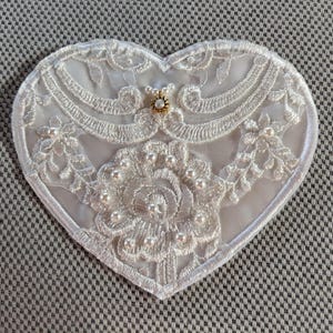 May include: White heart-shaped lace applique with a pearl and rhinestone embellishment. The lace is delicate and intricate, with a floral design.