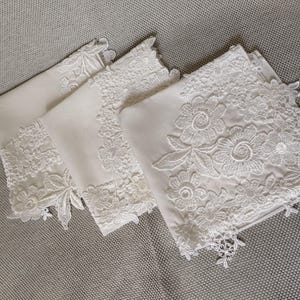 May include: Four white linen handkerchiefs with intricate floral lace detailing. The lace features various flower designs and scalloped edges. The handkerchiefs are neatly folded and arranged on a gray surface.