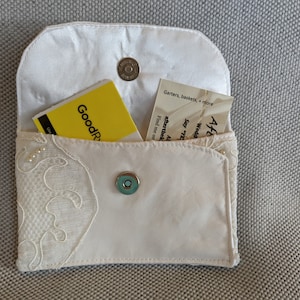 May include: A white satin pouch with a lace trim and a magnetic closure. The pouch is open and contains a yellow card with the text "GoodR" and a white card with the text "Garters, baskets, + more After the Wedding Say "Yes" to aftertheknot.com AL Find me on GoodR".