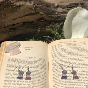 Puede incluir: Un par de pendientes de plata con un diseño a rayas azules y rojas. Los pendientes tienen forma de mujer que lleva una camisa a rayas y pantalones cortos rojos. Los pendientes cuelgan de un libro con el texto "The World Is Like That" en la página.