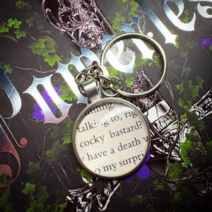 May include: A round silver keychain with a printed quote: "talking to, cocky bastard?" The keychain is attached to a silver chain and ring. The background is a dark, patterned surface with floral and gothic elements.