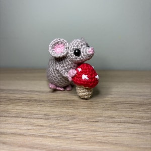 May include: A small, crocheted grey mouse figurine with pink inner ears and a pink nose. The mouse has black bead eyes and is holding a crocheted mushroom with a red cap featuring white embroidered spots and a light tan stem.