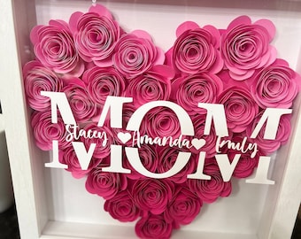 Könnte beinhalten: Ein weißer Rahmen mit einem rosa Papierblumenherz, auf dem "MOM" steht, und den Namen "Stacey", "Amanda" und "Emily" im Herzen.
