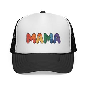 Puede incluir: Gorra de camionero en blanco y negro con la palabra "MAMA" en letras de rompecabezas arcoíris. La gorra tiene un panel frontal blanco, una visera negra y una parte trasera de malla negra.