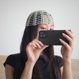 Könnte beinhalten: Ein hellgrüner Häkel-Beanie mit offenem Webmuster. Der Beanie wird auf dem Kopf getragen. Die Person hält ein schwarzes Smartphone.