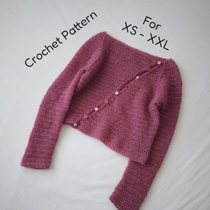 May include: A mauve crochet cardigan with long sleeves and a diagonal button closure featuring small white and pink buttons. The text Crochet Pattern For XS - XXL is visible above the garment, indicating the available sizing for this design.