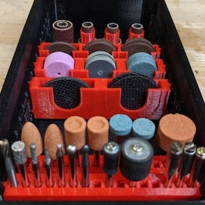 Puede incluir: Una caja organizadora negra y roja llena de varias brocas y accesorios para herramientas rotativas. La caja tiene múltiples compartimentos que contienen muelas, tambores de lijado y otras herramientas pequeñas. Las herramientas son de varios colores y formas.