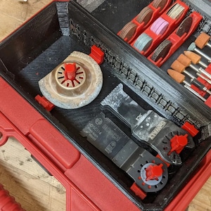 May include: A red tool case with a black interior. The case is open and contains a variety of tools, including a saw blade, a sanding disc, and a set of small drill bits. The tools are organized in a variety of compartments and slots.