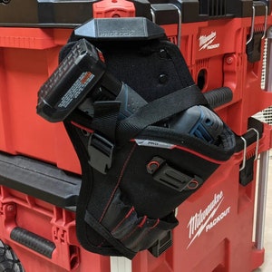 May include: A black tool belt with a red and black Milwaukee Packout logo. The belt is holding a black and blue cordless drill. The belt is attached to a red toolbox.