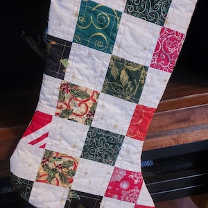 May include: A quilted Christmas stocking with a patchwork design. The stocking features squares of red, green, and white fabric with gold accents and festive patterns. It hangs from a wooden mantel.