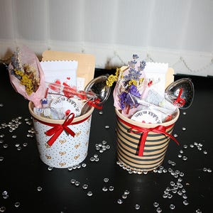 Könnte beinhalten: Zwei kleine Papierbecher gefüllt mit Geschenken. Jeder Becher hat ein rotes Band darum gebunden. Die Becher sind mit einem weißen und goldenen Tupfenmuster und einem schwarz-weißen Streifenmuster verziert. Die Becher enthalten getrocknete Blumen, Teebeutel, einen Tee-Infuser und eine kleine Karte.