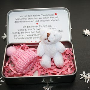 Könnte beinhalten: Eine kleine, offene Metalldose mit einem weißen Teddybären und einer rosa Strickmütze mit Bommel, eingebettet in rosa Papierschnitzel. Die Dose hat eine gedruckte Nachricht auf Deutsch. Dekorative Schneeflocken sind verstreut.