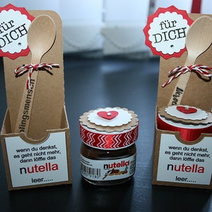 Könnte beinhalten: Zwei braune Pappkartons mit einem Holzlöffel und einem kleinen Glas Nutella. Die Kartons haben rote und weiße Akzente und den Text "für DICH" auf der Vorderseite. Der Text "wenn du denkst, es geht nicht mehr, dann löffle das nutella leer..." ist auf der Unterseite der Kartons gedruckt.