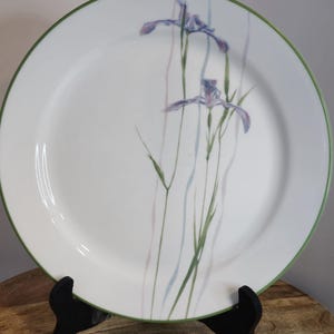 May include: A white ceramic dinner plate with a green rim and a floral design featuring purple irises and green leaves on a white background.
