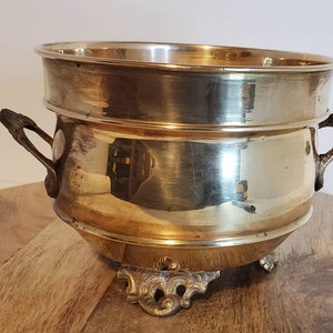 May include: A gold-toned brass planter with two handles and three decorative feet. The planter has a shiny, reflective surface.