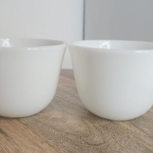May include: Two white ceramic mugs with rounded edges and a simple handle. The mugs are sitting on a light brown wooden surface.