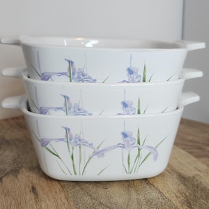 May include: Three white ceramic baking dishes with a floral design featuring purple irises and green leaves. The dishes are stacked on top of each other.