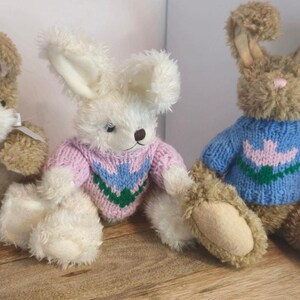 May include: Three plush bunny rabbits, each wearing a different colored knitted sweater. The first bunny is brown with white feet and pink ears, the second bunny is white with pink and green sweater, and the third bunny is brown with blue sweater and pink flower design.