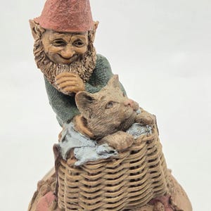 May include: A figurine of a gnome with a textured red hat and green shirt, holding a small cat inside a woven basket. The base features sculpted leaves and acorns with the text CATFISHING, Clark, and Wolf, along with a signature etched into the surface.
