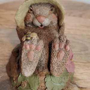 May include: A small, brown rabbit figurine wearing a green hat and sitting on a brown base with green leaves. The rabbit is holding a small, yellow and black bee figurine.
