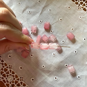 May include: Pink and white rectangular beads are strung on a gold wire, with additional loose beads scattered on a white, eyelet-embroidered cloth. The beads have a translucent quality, and the color gradient varies from pink to white.