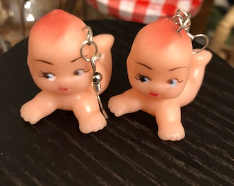 Op de afbeelding: Een paar oorbellen met kleine, roze plastic babypoppen met gesloten ogen en geverfde wimpers. De poppen zijn in een kruipende positie met uitgestrekte armen en benen. De oorbellen zijn gemaakt met zilverkleurige metalen haakjes.