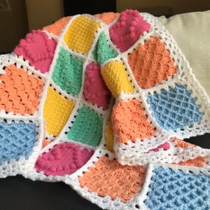 May include: A colorful crocheted baby blanket with a white border. The blanket is made up of squares in different colors, including pink, blue, green, yellow, and orange. The squares have a textured, bumpy surface.