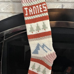 May include: A crocheted Christmas stocking with a pink, white, green, and orange color scheme. The stocking has a name "JAMES" embroidered on the top and a pattern of trees and mountains.