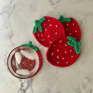 May include: Three crocheted strawberry-shaped coasters in red with green leaves. The coasters are on a white marble surface.