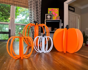 May include: Four decorative pumpkins, two orange and two white, are arranged on a wooden table. The pumpkins are made of layered circles and have a three-dimensional appearance.