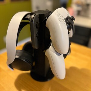 May include: A black stand with a white headset and two white video game controllers. The stand has a black base and a black column with a white headset attached to the top. The two white controllers are attached to the column, one on each side.