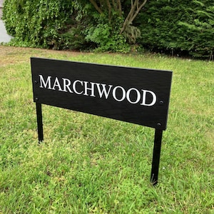 Puede incluir: Un letrero rectangular negro con la palabra MARCHWOOD impresa en letras mayúsculas blancas. El letrero está montado sobre dos estacas de metal negro delgadas insertadas en el suelo. La superficie tiene una textura de veta de madera horizontal.