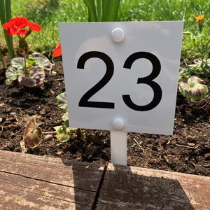 Könnte beinhalten: Ein quadratisches weißes Gartenschild mit der schwarzen Zahl 23 in einer fetten serifenlosen Schrift. Das Schild ist auf einem weißen Pfahl montiert und mit zwei weißen runden Befestigungselementen in der oberen und unteren Mitte befestigt.