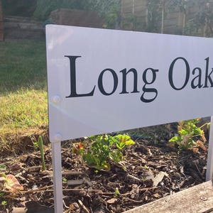 May include: A white sign with the words "Long Oak" in black, mounted on white posts in a garden. The garden bed features pink flowers and green foliage, with a wooden border.