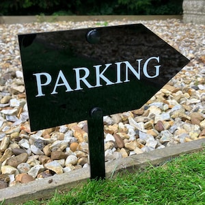 Pode incluir: Placa preta em forma de seta com texto branco que diz "PARKING" em uma estaca de metal preta. A placa está colocada em um leito de cascalho.