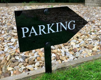 May include: Black arrow sign with white text that reads "PARKING" on a black metal stake. The sign is placed in a bed of gravel.