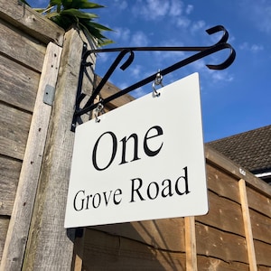May include: White sign with black text that reads "One Grove Road" hanging from a black metal bracket attached to a wooden fence.