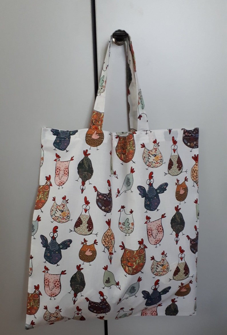 May include: A white tote bag with a colorful pattern of chickens. The chickens are drawn in a cartoon style and have different colors and patterns. The bag has two long handles.