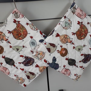 Könnte beinhalten: Zwei weiße, gesteppte Topflappen mit einem Muster aus bunten Hühnern. Die Topflappen haben oben eine Schlaufe zum Aufhängen.