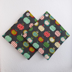 Könnte beinhalten: Zwei quadratische Topflappen mit dunkelgrauem Hintergrund und einem sich wiederholenden Muster aus bunten Äpfeln. Die Äpfel weisen verschiedene Designs auf, darunter Streifen, Tupfen und Volltonfarben. Jeder Topflappen hat eine Metallöse in einer Ecke.