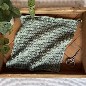 Op de afbeelding: Een saliegroene, gehaakte vierkante deken met een textuurpatroon, rustend op een houten dienblad met houten kralen handvatten. Een kleine schaar en een potplant staan ook op de dienblad.