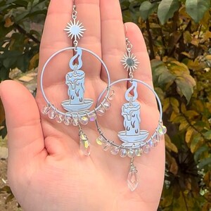 May include: A pair of silver hoop earrings with a candle and star charm. The candle is blue and white and the star is silver. The earrings are decorated with clear iridescent beads.