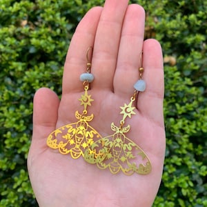 May include: Gold tone earrings featuring a detailed cutout of a moth with celestial elements, including stars, moons, and a crescent moon. The earrings are accented with a light blue stone.