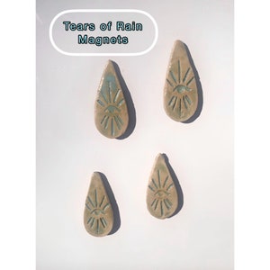 May include: Four teardrop-shaped magnets with an eye design and sunburst rays. The magnets are beige with turquoise accents and are arranged on a white surface. The text "Tears of Rain Magnets" is in a white oval.
