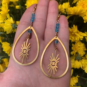 May include: Gold teardrop earrings with a sunburst design and a blue eye in the center. The earrings are accented with blue crystal beads.