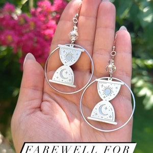 May include: A pair of silver hoop earrings with a sun and moon hourglass charm. The earrings are hanging from a hand.